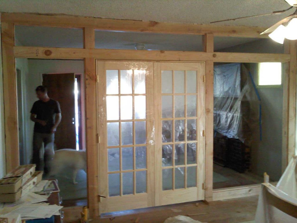 A room undergoing renovation with wooden work in progress. A man is standing in the room, inspecting the renovation work. Tools and materials are scattered around the room, indicating ongoing construction